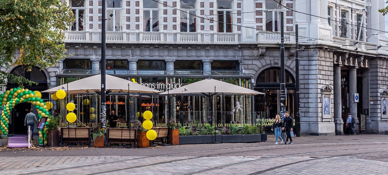 Een straatbeeld toont een café met grote ramen en zitplaatsen buiten onder bruine parasols, versierd met gele ballonnen. Verschillende mensen lopen voorbij en het gebouw erachter heeft een sierlijke gevel met stenen bogen.
