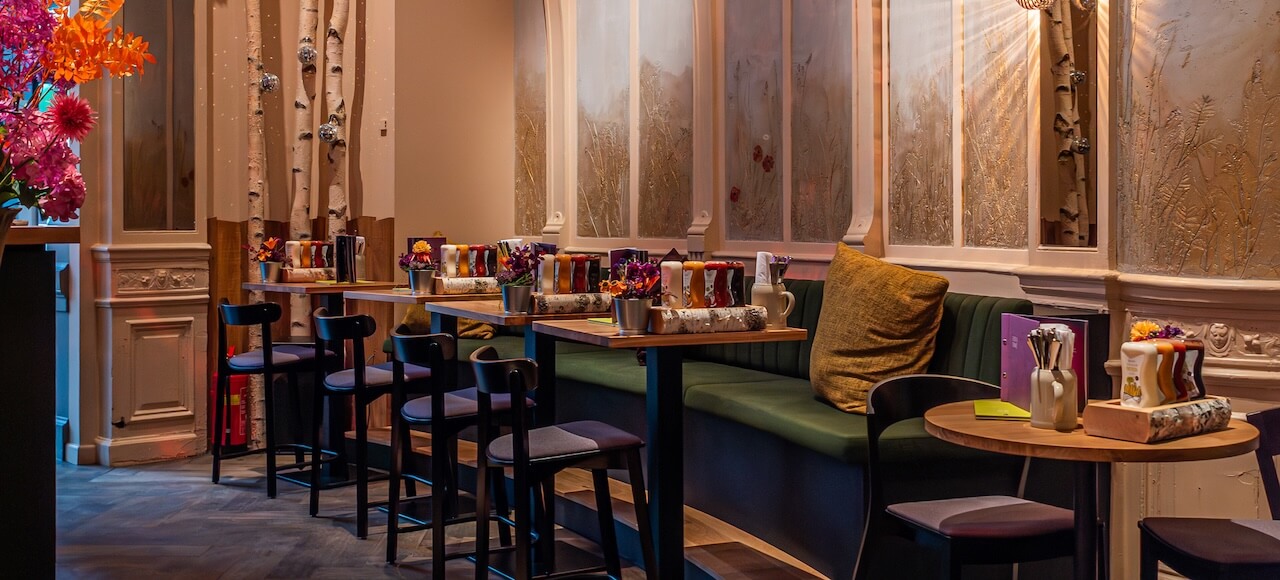 Een gezellig café met donkerhouten tafels en stoelen, groene banken met kussens, vazen met kleurrijke bloemen en zachte verlichting. Decoratieve wandpanelen en accenten van berkenbomen creëren een warme en uitnodigende sfeer.