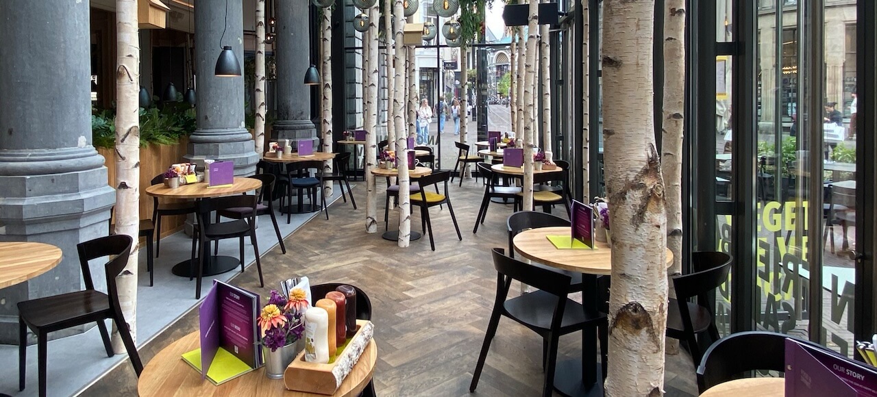 Een modern restaurantinterieur met ronde houten tafels, zwarte stoelen, hoge zuilen, berkenstammen en grote ramen. De tafels zijn gedekt met menu's, specerijen en bloemen; buiten op straat zijn mensen te zien.
