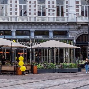 Een straatbeeld toont een café met grote ramen en zitplaatsen buiten onder bruine parasols, versierd met gele ballonnen. Verschillende mensen lopen voorbij en het gebouw erachter heeft een sierlijke gevel met stenen bogen.