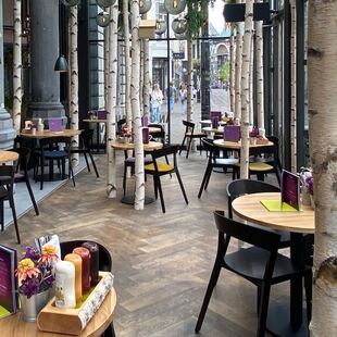 Een modern restaurantinterieur met ronde houten tafels, zwarte stoelen, hoge zuilen, berkenstammen en grote ramen. De tafels zijn gedekt met menu's, specerijen en bloemen; buiten op straat zijn mensen te zien.