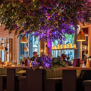 Een stijlvol restaurantinterieur met een grote boom als middelpunt, kleurrijke verlichting, elegante inrichting, pluche groene stoelen, gouden hanglampen en levendige bloemstukken, met ramen die het groen buiten onthullen.