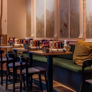 Een gezellig café met donkerhouten tafels en stoelen, groene banken met kussens, vazen met kleurrijke bloemen en zachte verlichting. Decoratieve wandpanelen en accenten van berkenbomen creëren een warme en uitnodigende sfeer.